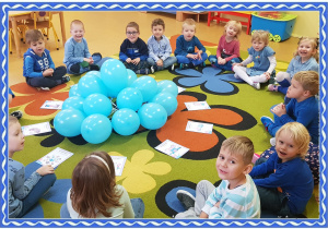 Grupa dzieci siedzi na dywanie. przed nimi leżą niebieskie balony i obrazki przedstawiające prawa dziecka.